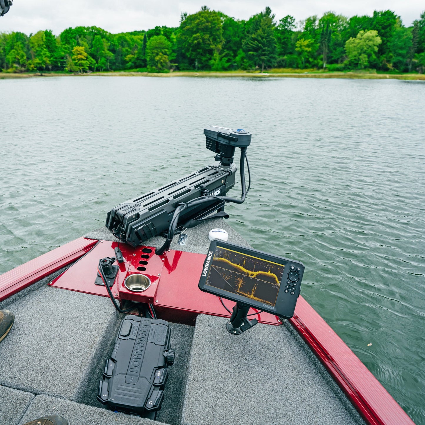 Person on a boat with fishing equipment and screen displaying sonar data over a lake.
