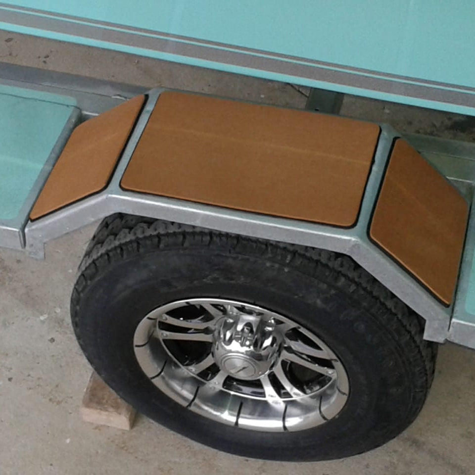 Close-up of a boat trailer wheel with a black tire and chrome rim, set against a silver metal frame with tan protective panels and light aqua side walls.