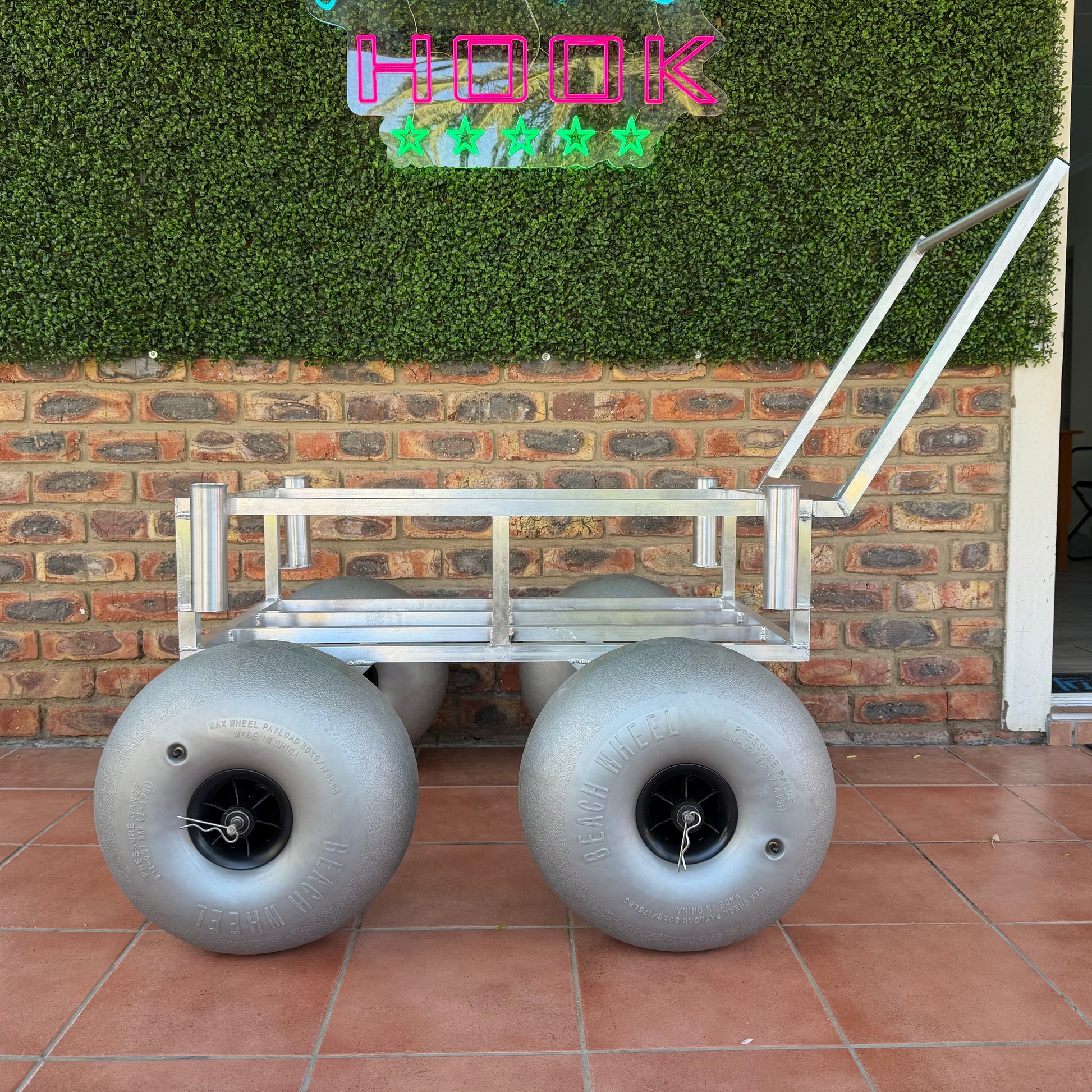 Metal cart with large wheels on a tiled patio, against a brick wall with greenery.