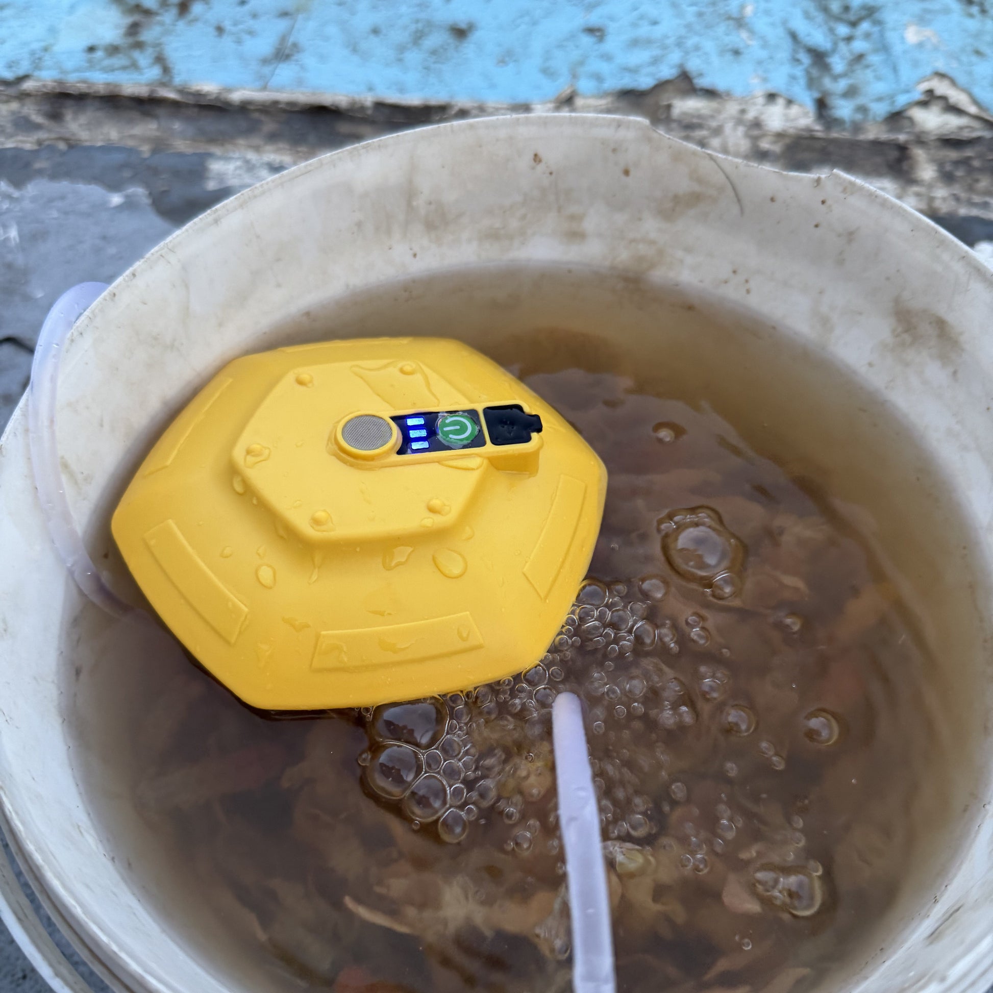 A yellow underwater water quality monitor with a small digital display, floating in a bucket of murky brown water with a bubble tube extending downward.