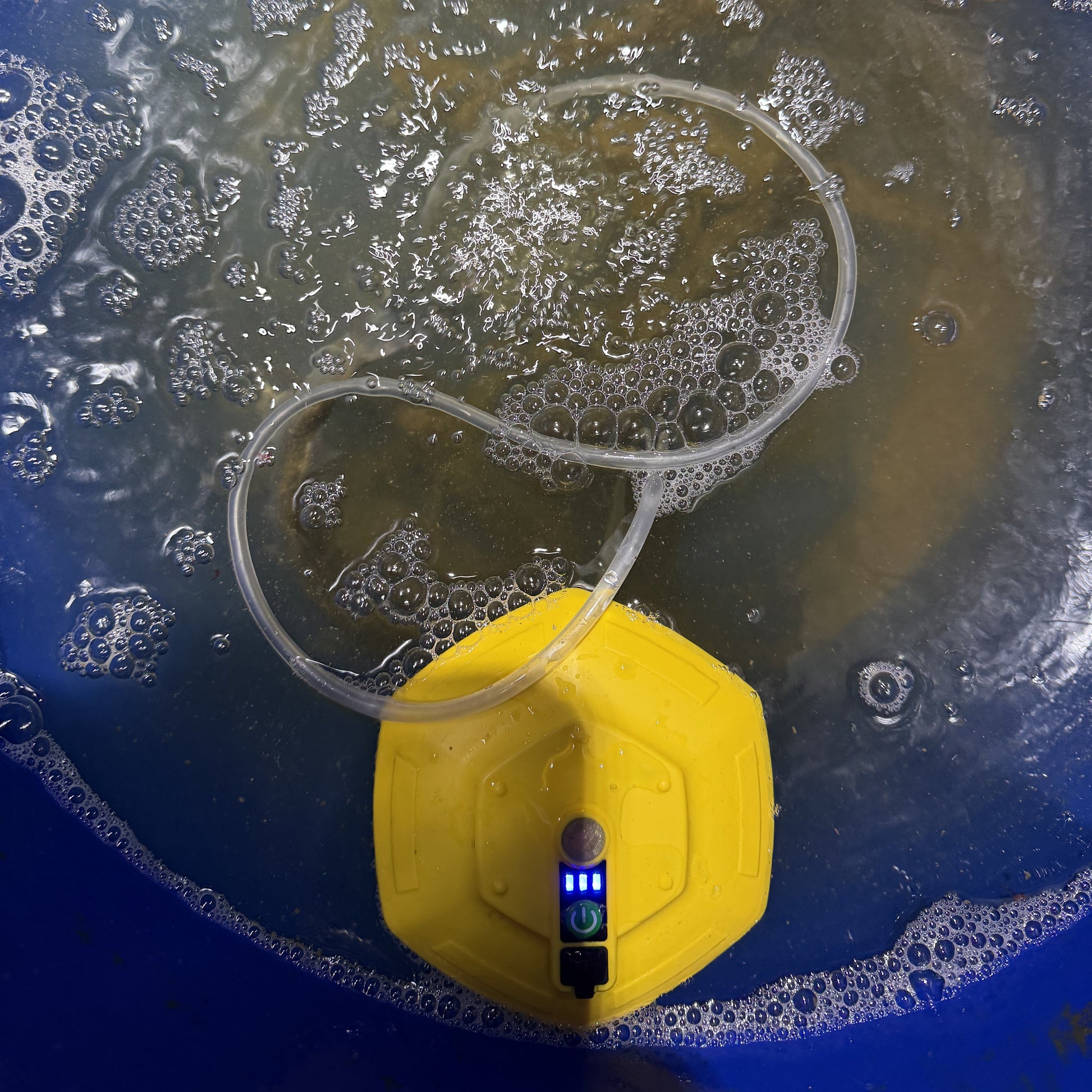 A yellow underwater bubble stone aerator pump with a clear tubing loop, shown inside a blue bucket with bubbling water and a fish swimming above.