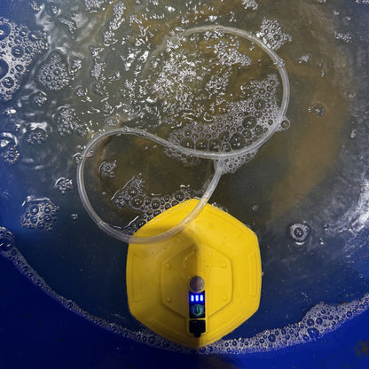 A yellow underwater bubble stone aerator pump with a clear tubing loop, shown inside a blue bucket with bubbling water and a fish swimming above.