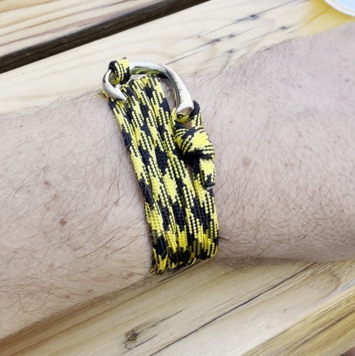 A close-up of a person's wrist wearing a black and yellow paracord bracelet with a silver metal carabiner clasp, resting on a light wood surface.