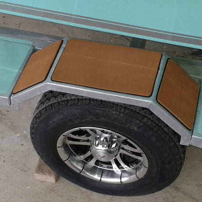 Close-up of a boat trailer wheel with a black tire and chrome rim, set against a silver metal frame with tan protective panels and light aqua side walls.