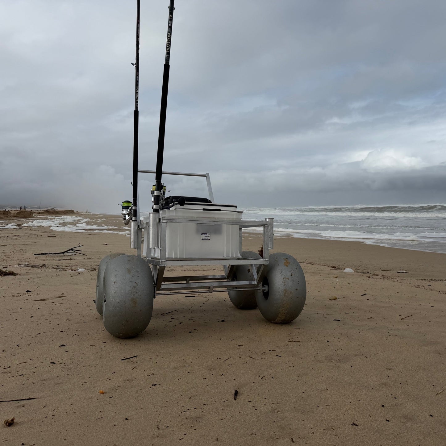 Tidal Hook beach fishing cart with oversized balloon wheels, storage crate, and rod holders positioned on the shoreline for easy transport of surf fishing equipment.