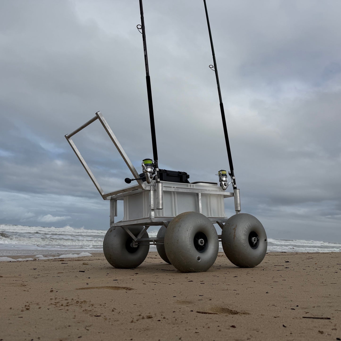 Beach fishing cart with oversized balloon wheels, dual rod holders and storage crate shown on soft sand for easy transport of surf fishing gear.