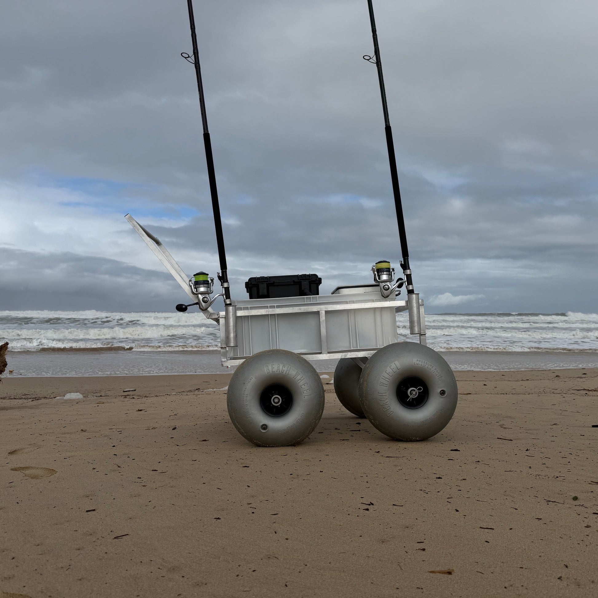Heavy-duty beach fishing cart with oversized balloon wheels, rod holders and storage crate designed for transporting surf fishing gear across soft sand.