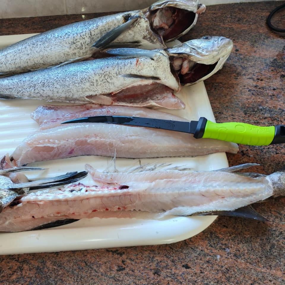 Black fillet knife with bright green handle being used to fillet freshly caught fish on a cleaning table.