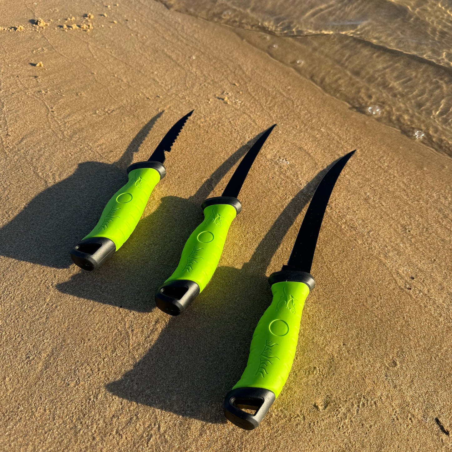 Three black fillet knives with bright green handles displayed on a sandy beach, showcasing different blade styles for fish cleaning and filleting.