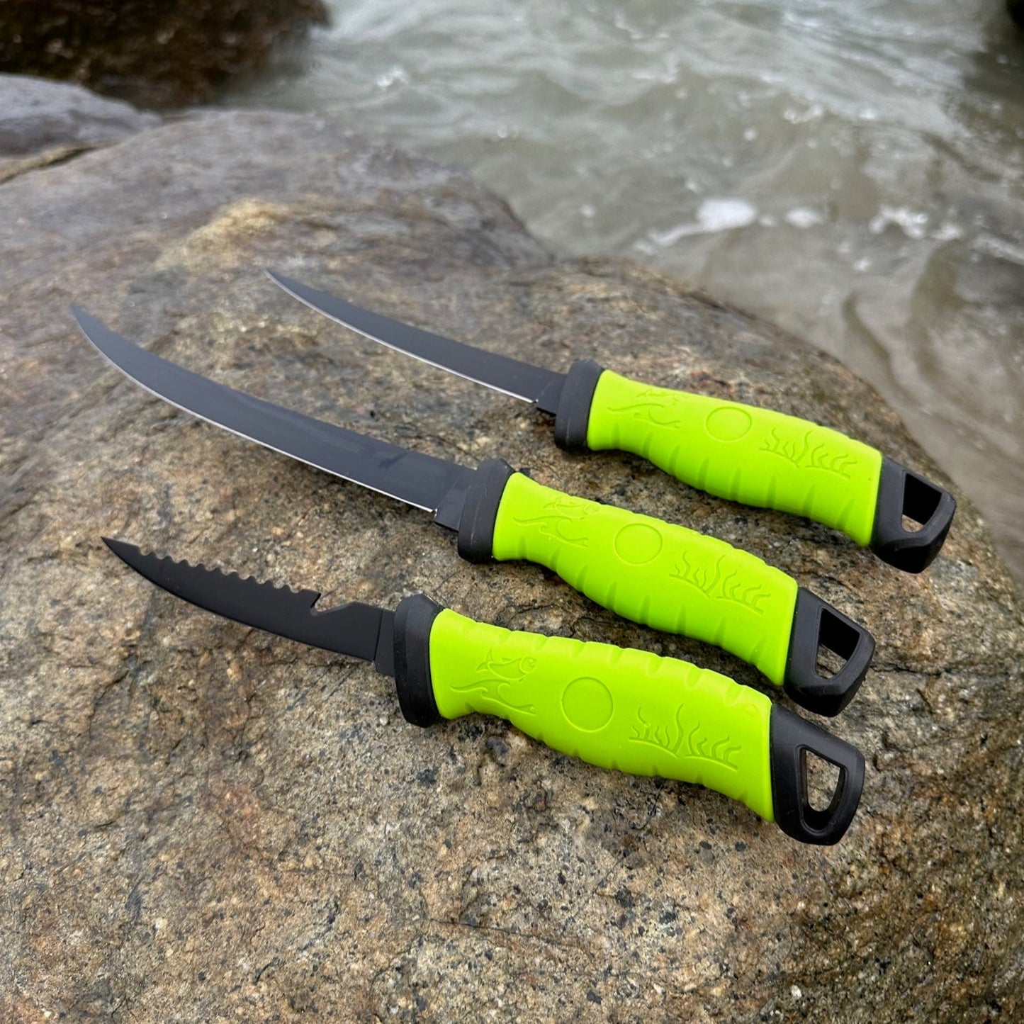 Three black fillet knives with bright green handles displayed on a rock beside the water, showcasing multiple blade styles for fish cleaning and filleting.