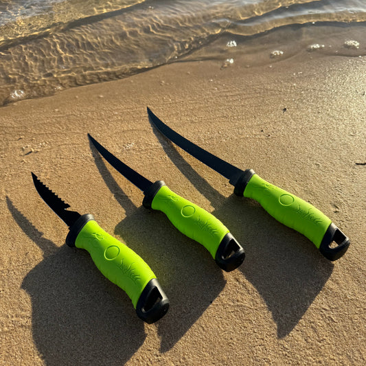 Three black fillet knives with bright green handles displayed on a sandy beach, designed for precision fish cleaning and filleting.